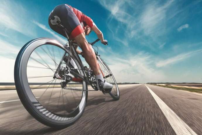 An athlete is riding a bicycle on road. The man is wearing black bike shorts and shin guards along with a red sleeveless top and a red and white helmet and sunglasses. The image is blurred in motion.
