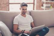 Young attractive smiling guy is browsing at his laptop, sitting at home on the cozy beige sofa at home, wearing casual outfit