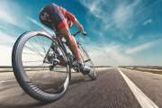 An athlete is riding a bicycle on road. The man is wearing black bike shorts and shin guards along with a red sleeveless top and a red and white helmet and sunglasses. The image is blurred in motion.