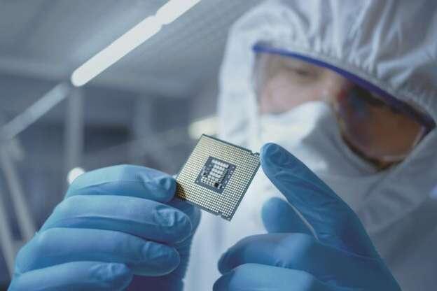 In Ultra Modern Electronic Manufacturing Factory Design Engineer in Sterile Coverall Holds Microchip with Gloves and Examines it.