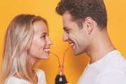 Pretty young couple standing isolated over yellow background, drinking fizzy drink from a glass bottle, looking at each other