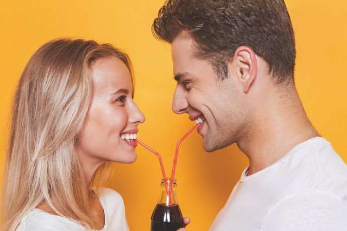 Pretty young couple standing isolated over yellow background, drinking fizzy drink from a glass bottle, looking at each other