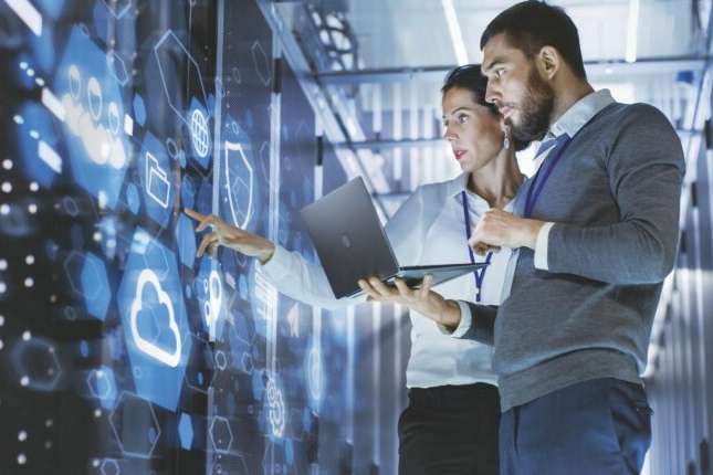 Male IT Specialist Holds Laptop and Discusses Work with Female Server Technician. They're Standing in Data Center, Rack Server Cabinet with Cloud Server Icon and Visualization.