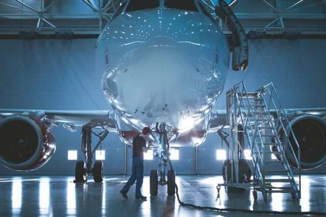 Brand New Airplane Standing in a Aircraft Maintenance Hangar while Aircraft Maintenance Engineer/ Technician/ Mechanic goes inside Cabin via Ladder/ Ramp.
